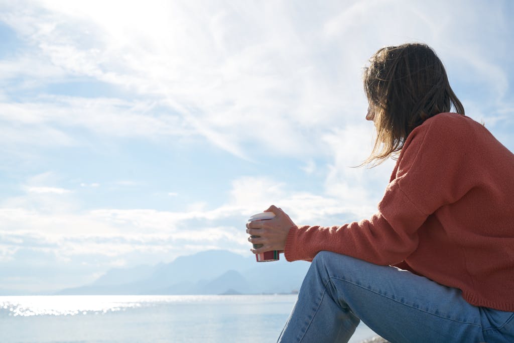 Eine Frau in Freizeitkleidung genießt ein heißes Getränk, während sie auf das Meer blickt – ein Inbegriff der Entspannung. Auch, wenn man mal keine Zeit hat für Gesangsübungen, ist das okay.